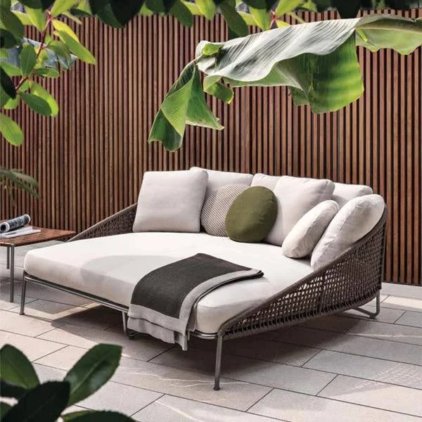 “Patio rope daybed set in dark brown frame with cream cushions, placed outdoors on a wooden deck surrounded by greenery.”