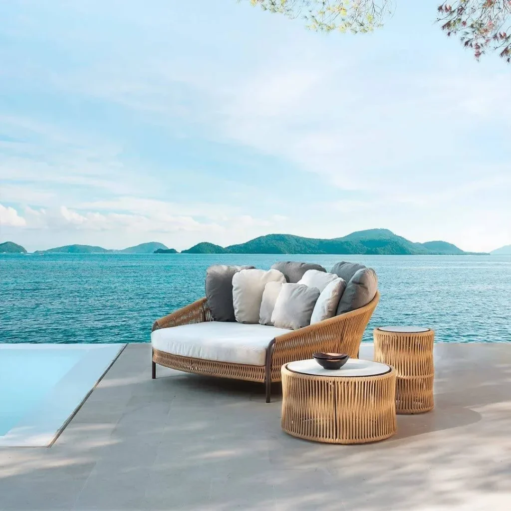 Beige and off-white outdoor rope daybed with plush cushions on a wooden deck surrounded by greenery.