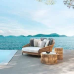 Beige and off-white outdoor rope daybed with plush cushions on a wooden deck surrounded by greenery.