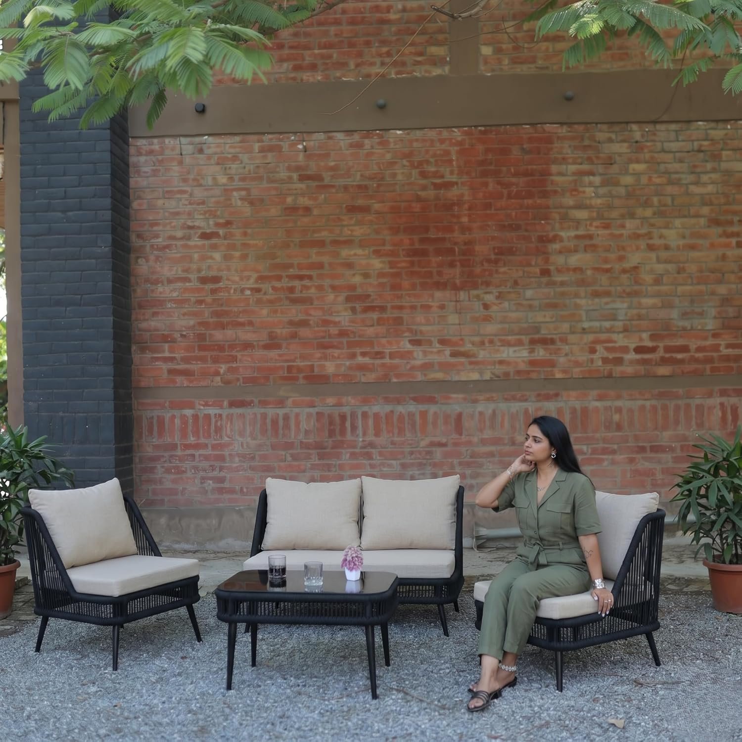 Outdoor patio furniture setup with a black woven sofa set, including a two-seater, two single chairs, and a coffee table with drinks and a small flower vase, placed on a gravel surface against a red brick wall. A woman in a green outfit is seated on one chair, relaxing under leafy trees in a garden setting.